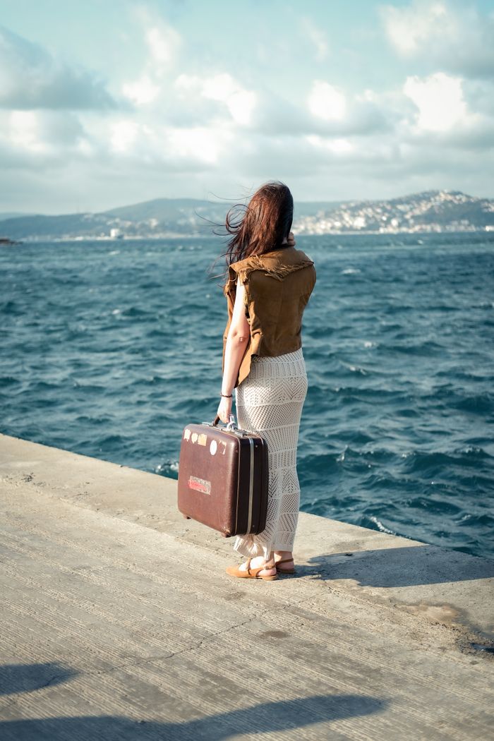 A woman stands on a beach promenade holding a vintage suitcase by the sea.
