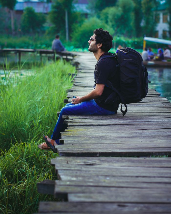 Adult man sitting on a wooden boardwalk by wetlands with a backpack, enjoying a peaceful moment.